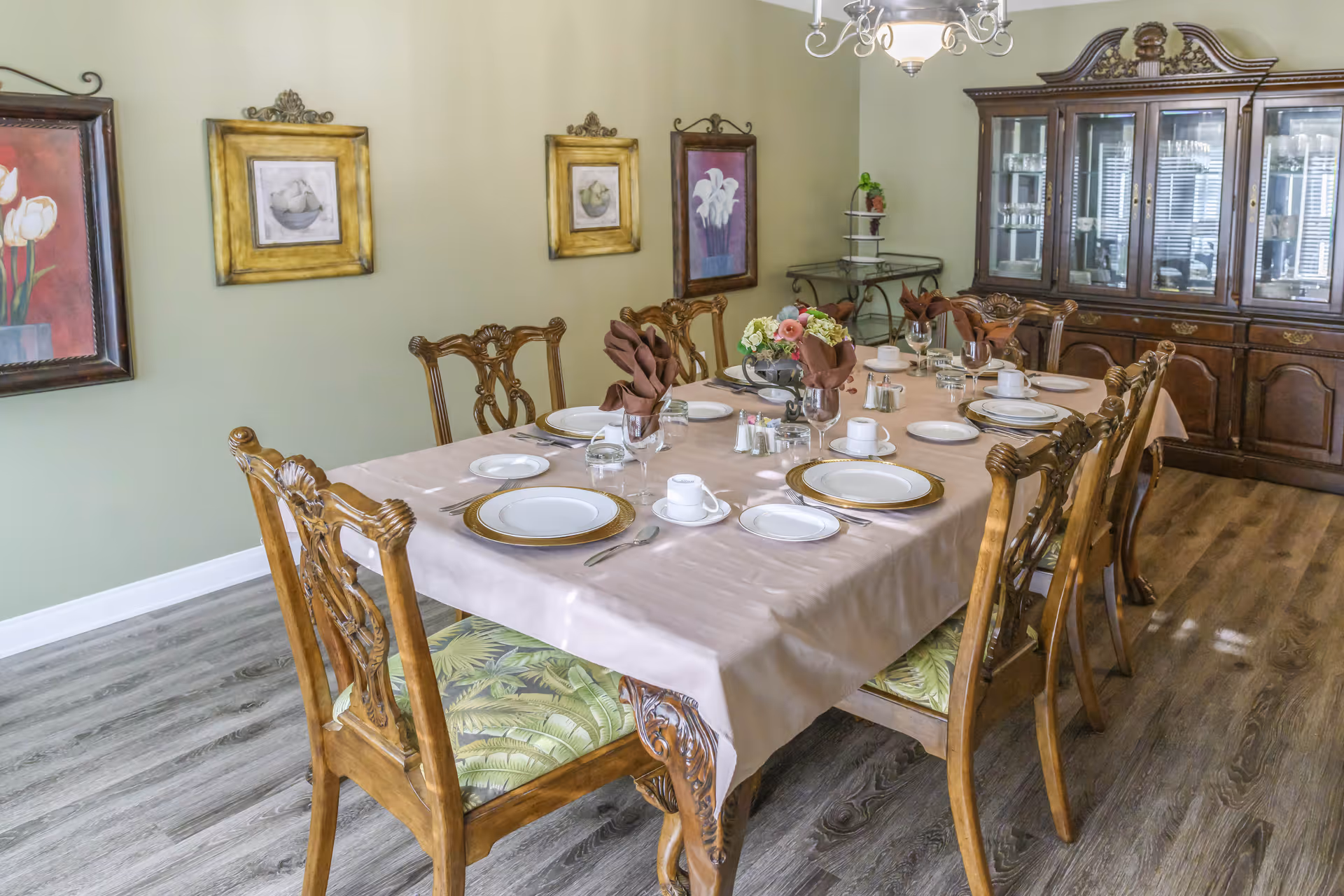 A formal dining room with a long wooden table covered with a beige tablecloth, set with plates, cups, glasses, and brown folded napkins. The room has wooden chairs with patterned cushions, a wooden china cabinet filled with glassware, framed floral artwork on the light green walls, and a chandelier hanging from the ceiling.