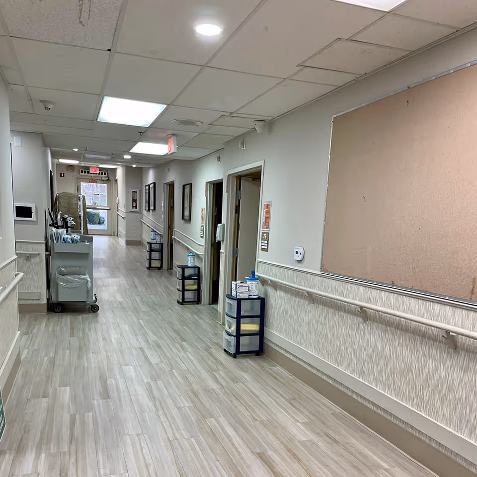 A clean, well-lit hallway in a senior living facility with light wood flooring, handrails on both sides, several open doorways, a bulletin board on the right wall, and a cart with supplies on the left side near the end of the hallway.