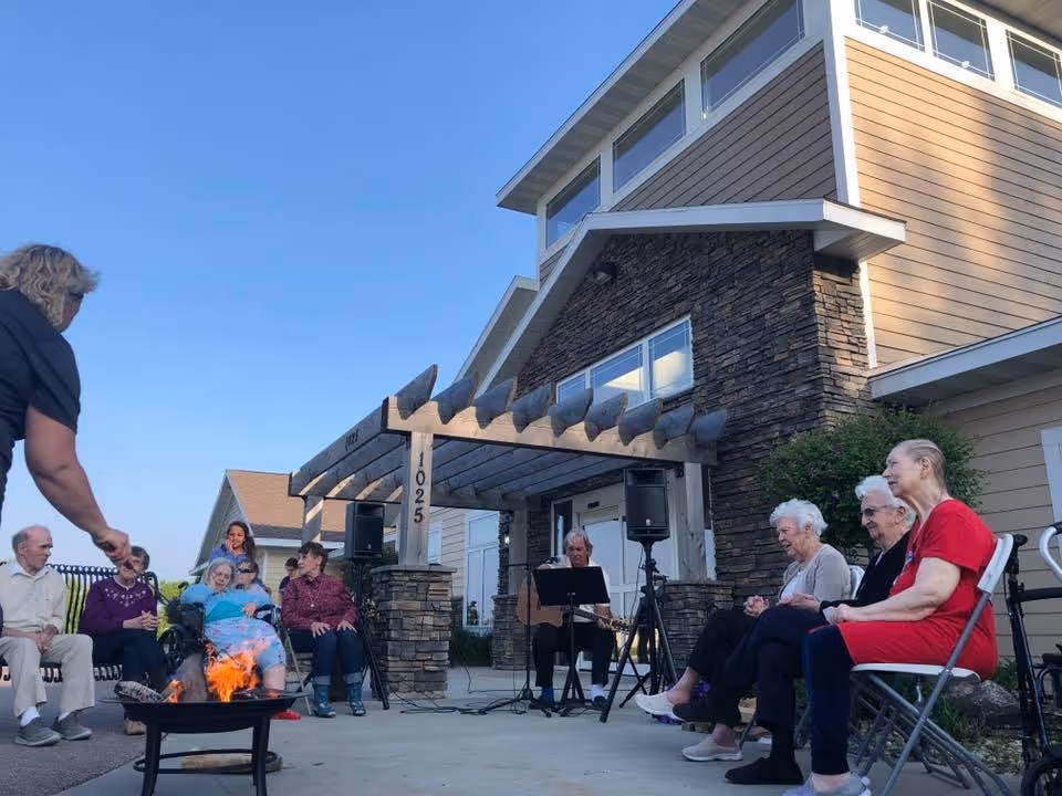 A group of elderly people sitting outdoors in front of a senior living facility building, enjoying a small fire pit and live music played by a man with a guitar under a wooden pergola.