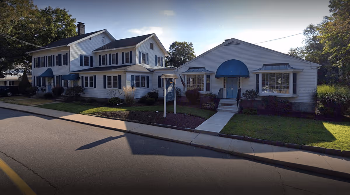 Exterior view of Essex Village Manor, showing two connected white buildings with blue awnings over the doors and windows, surrounded by a small lawn and sidewalk along a street.