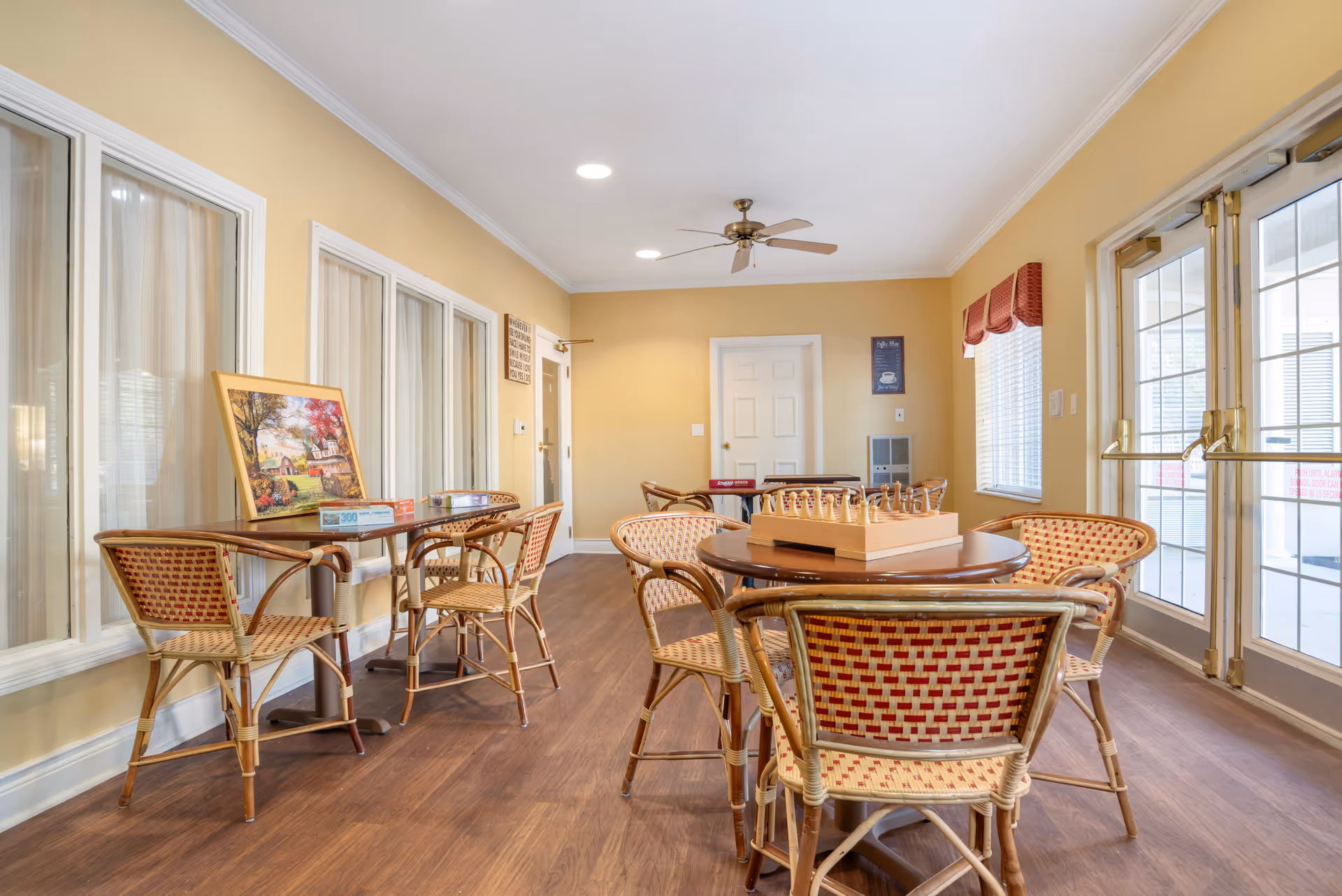 A bright room with yellow walls and wooden flooring featuring several round tables with wicker chairs arranged around them. One table has a chess set on it, and another table has a puzzle box and a framed picture resting on it. The room has large windows with white curtains on one side and glass double doors with red valances on the other side. A ceiling fan with lights is mounted on the white ceiling.
