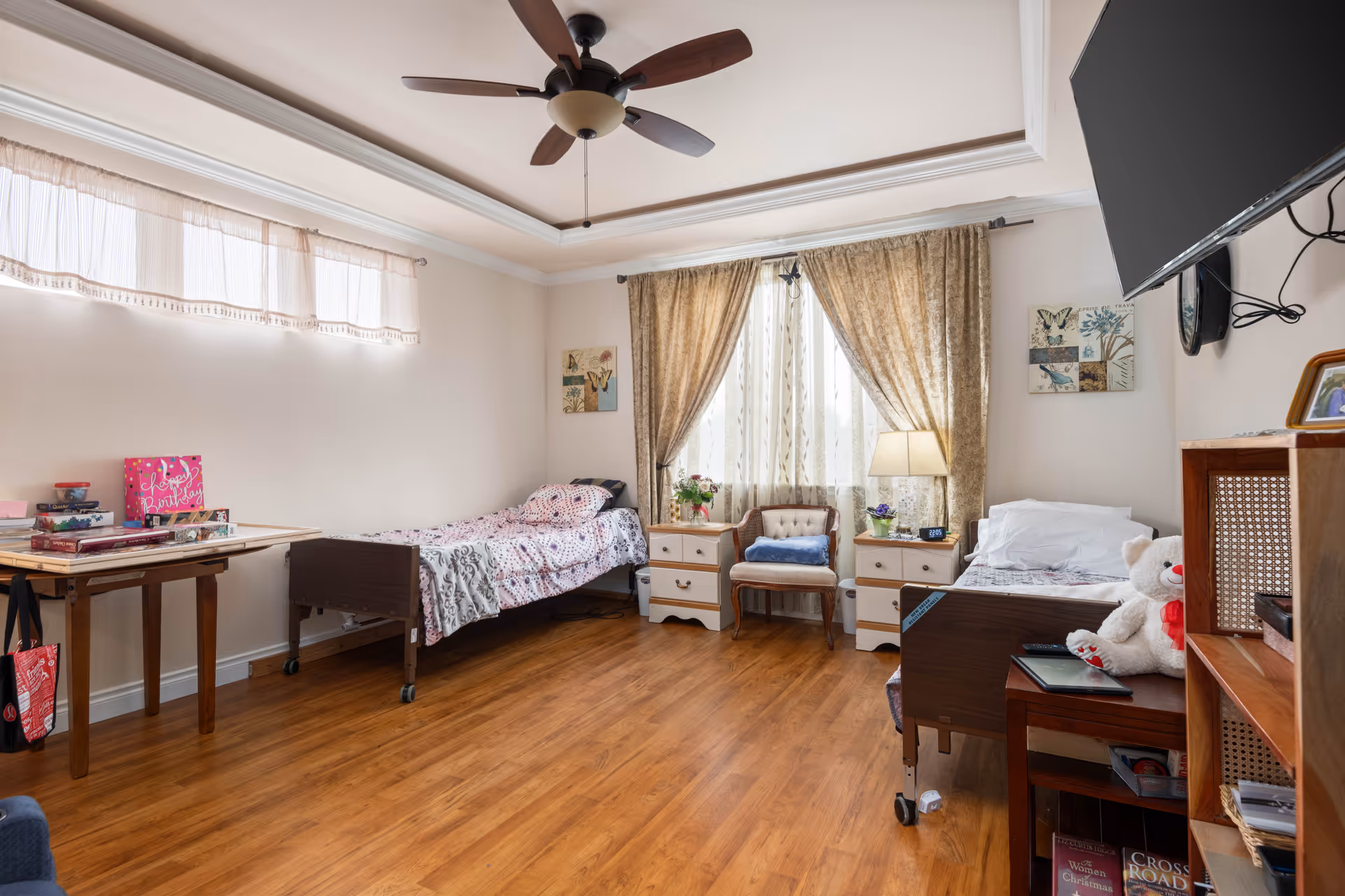 A bright and cozy bedroom with two single beds on opposite sides of the room, each with patterned bedding. Between the beds is a window with sheer and heavy curtains, flanked by two white nightstands with a lamp and plants. A wooden chair with a cushion sits in front of the window. The room has wooden flooring, a ceiling fan, a wall-mounted TV, and a wooden bookshelf with books and a teddy bear on one of the beds.