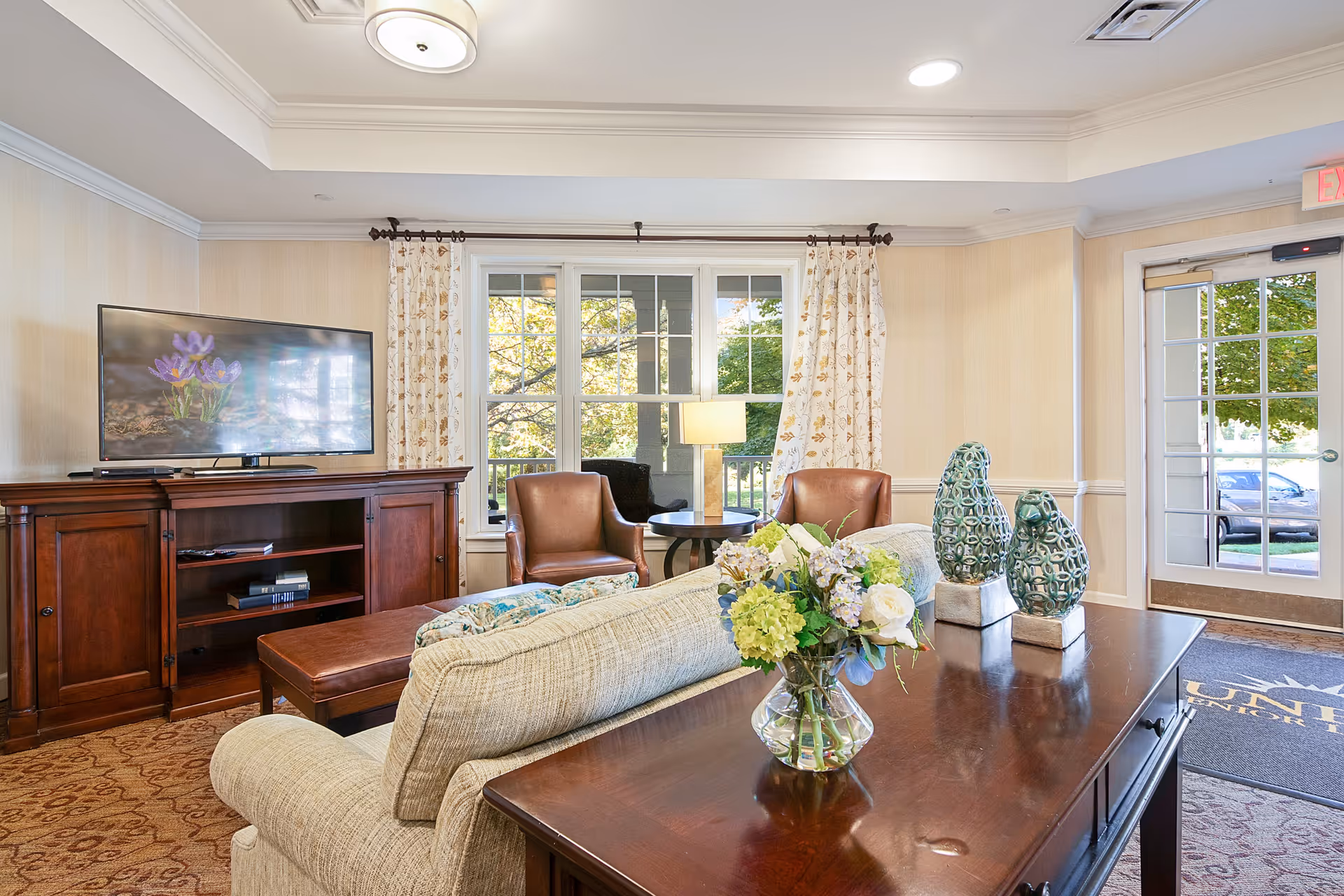 A cozy living room area in a senior living facility with a beige sofa, a wooden coffee table with decorative vases and a flower arrangement, two brown leather chairs, a wooden TV stand with a flat-screen TV displaying purple flowers, large windows with floral curtains, and a glass door leading outside.
