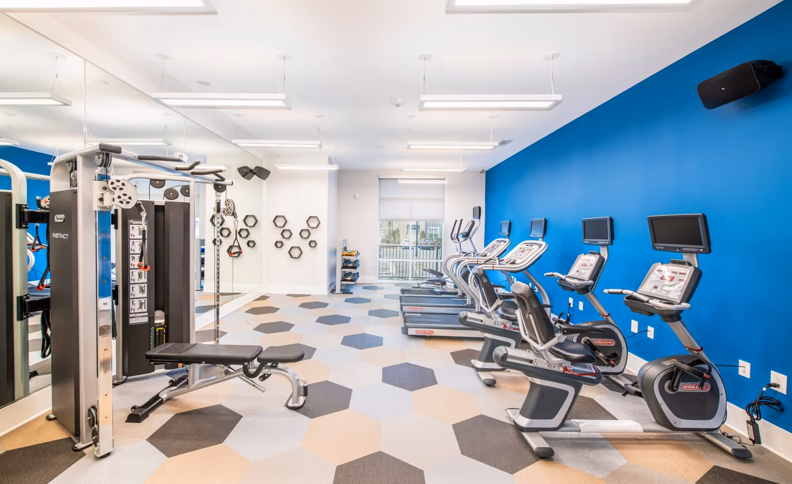 Bright fitness room with exercise equipment including treadmills, stationary bikes, and a multi-function weight machine. The room features a blue accent wall, large mirrors on one side, hexagonal patterned flooring, and modern ceiling lights.