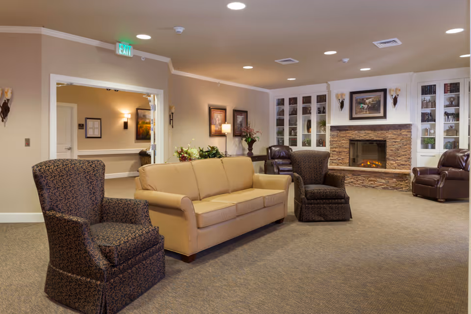 A cozy living room area in an assisted living facility featuring a beige sofa, two patterned armchairs, and two brown leather recliners. The room has a stone fireplace with a framed painting above it, built-in white shelves with decorative items, and warm lighting from wall sconces and ceiling lights. There is an open doorway leading to another room with framed artwork on the walls.
