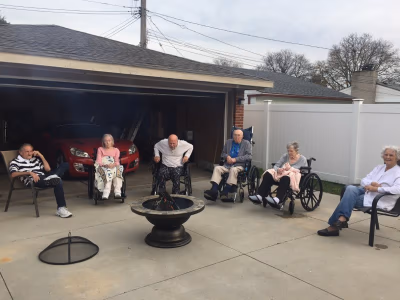 Six elderly individuals, some in wheelchairs and others seated in chairs, gathered around a fire pit on a concrete driveway in front of a garage with a red car inside. There is a white fence and leafless trees in the background under a cloudy sky.