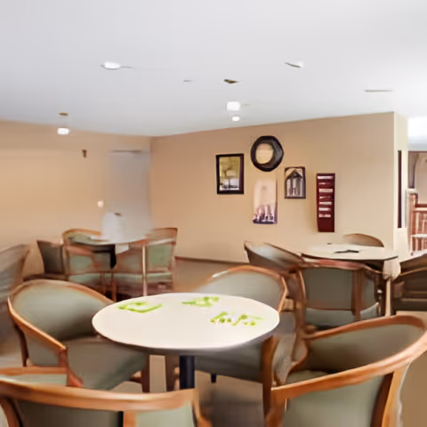 Communal dining/activity room with round tables and green-upholstered chairs and framed artwork on the wall.