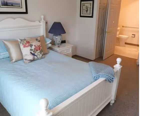 A bedroom with a white wooden bed frame and a light blue quilted bedspread. There are decorative pillows on the bed, including one with a floral and bird design. A white nightstand with a blue lamp is next to the bed. The room has beige walls and carpeted floor. An open door reveals a bathroom with a toilet and sink.