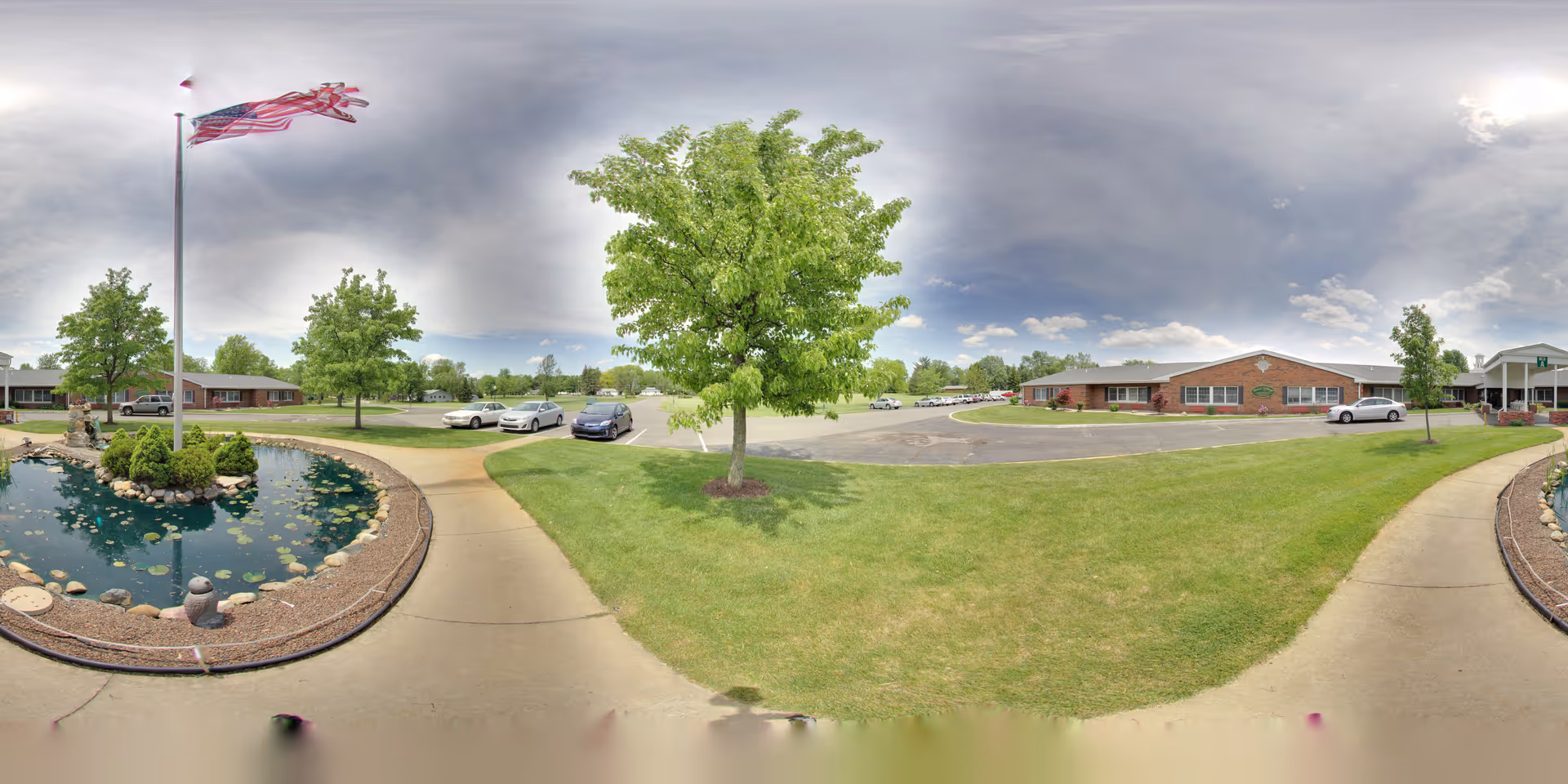 Wide panoramic view of the outdoor grounds at Grace Village Retirement Community showing a circular pond with rocks and plants, an American flag on a tall flagpole, green grass, several trees, a parking lot with cars, and single-story brick buildings under a partly cloudy sky.