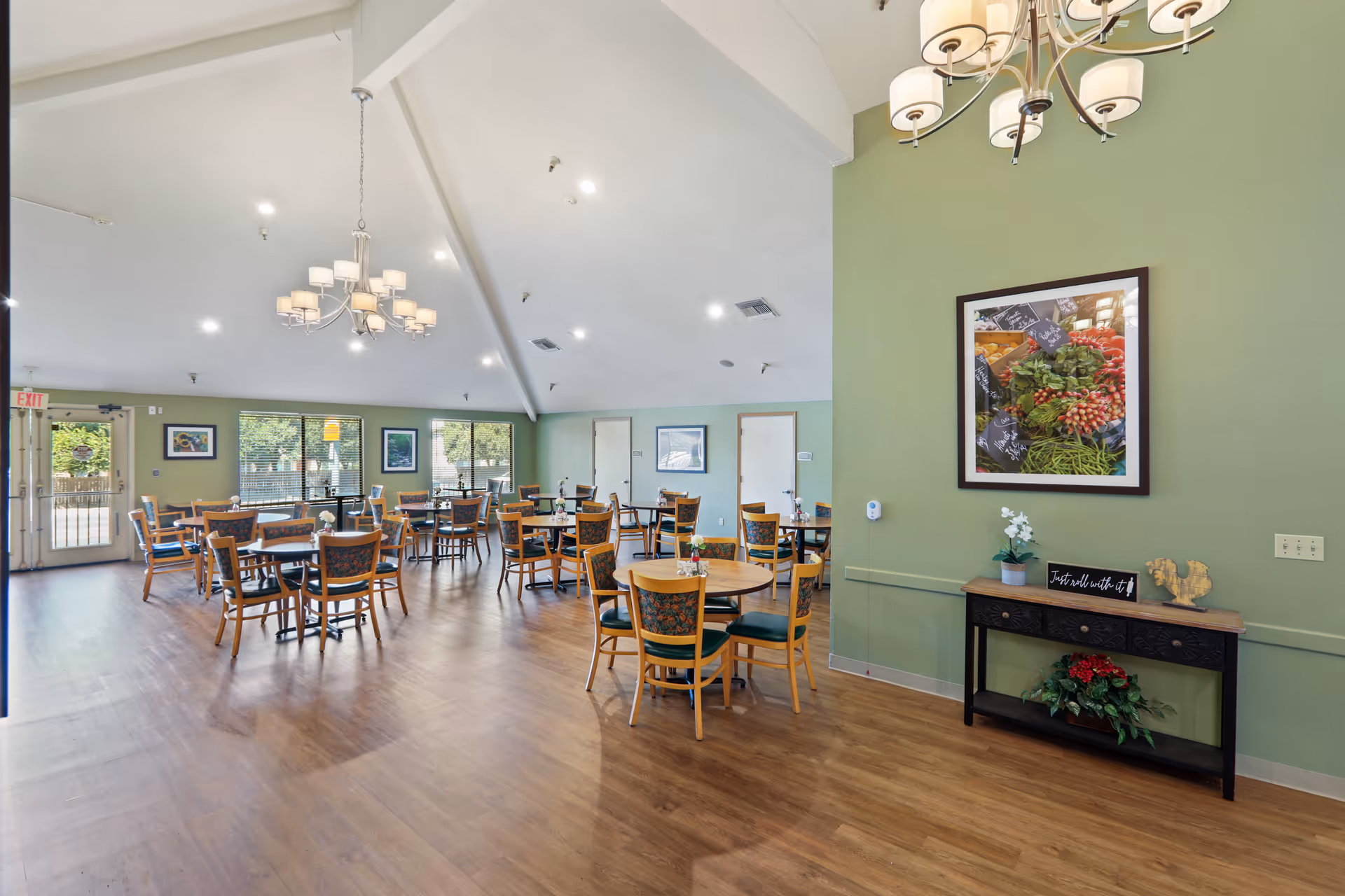 A spacious dining room with multiple round and square wooden tables surrounded by chairs with floral upholstery. The room has green walls, large windows letting in natural light, a wooden floor, and two modern chandeliers hanging from a vaulted ceiling. A console table with decorative items and a framed picture of fresh vegetables is against one wall.