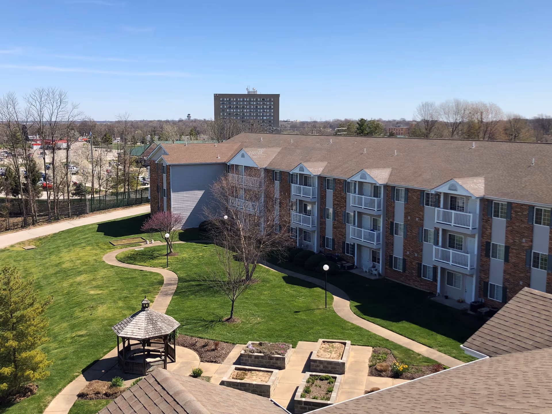 View of a senior living facility courtyard with a well-maintained lawn, a winding concrete pathway, a wooden gazebo, and several raised garden beds. The building is a multi-story brick and siding structure with balconies and windows. Trees and a clear blue sky are visible in the background.