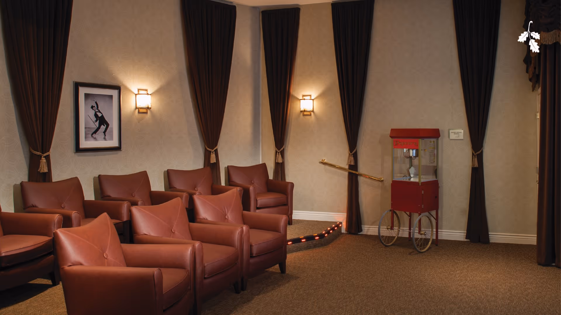 A cozy room with six brown leather armchairs arranged in two rows facing a popcorn machine on wheels. The walls are decorated with patterned wallpaper, three tall dark brown curtains, two wall sconces emitting warm light, and a framed black and white photo of a person dancing. There is a small step with red lights and a brass handrail leading to a door labeled 'Audio Visual Room'.