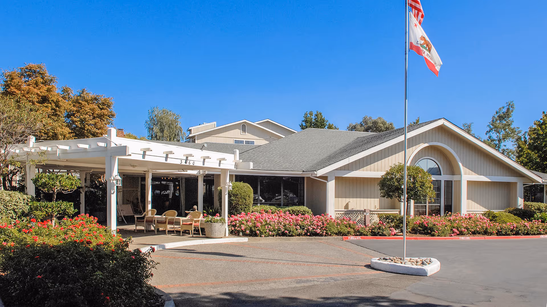 Exterior view of a single-story building with a gray roof and beige siding, surrounded by well-maintained landscaping including bushes and flowers. There is a white pergola with outdoor seating under it, and a flagpole with the American and California state flags in front of the building. The sky is clear and blue.