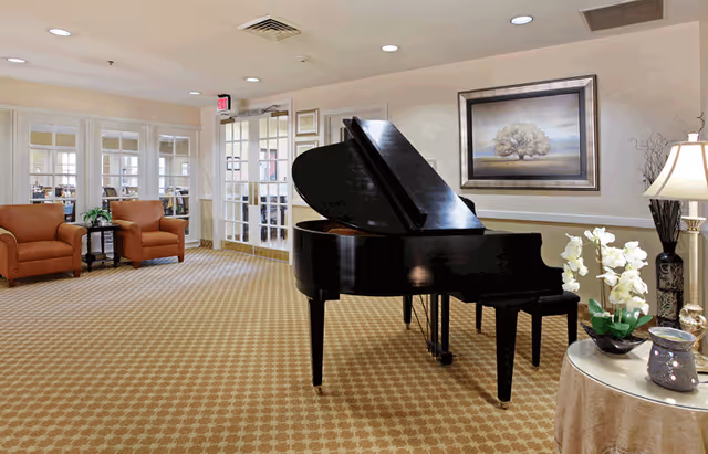 A spacious and well-lit common area featuring a black grand piano at the center. To the left, there are two orange armchairs with a small table between them, and a glass door leading to another room. On the right, a round table with a beige tablecloth holds a lamp, white orchids, and decorative items. A framed painting of a tree hangs on the wall behind the piano.