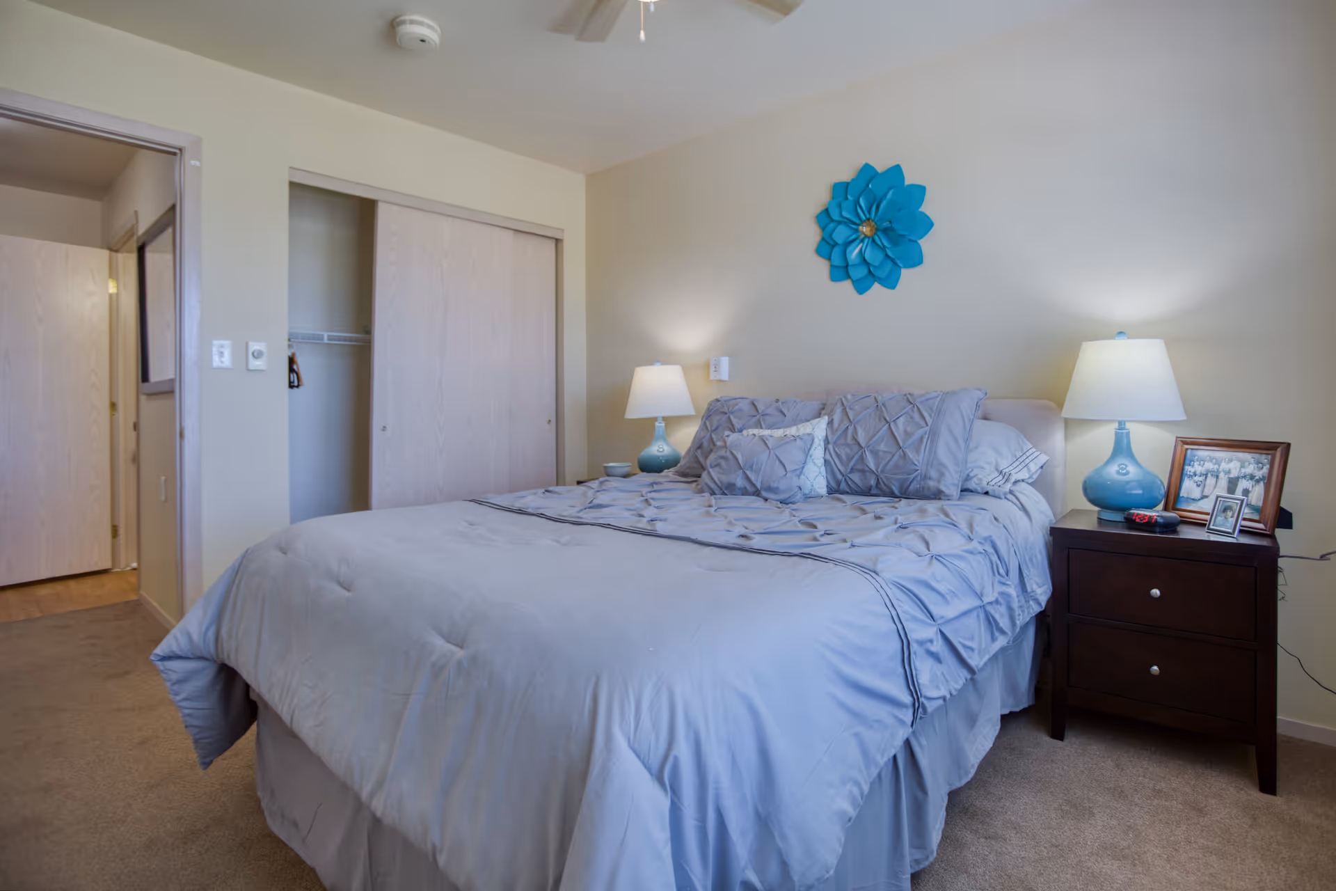 A bedroom with a neatly made bed featuring light blue bedding and multiple pillows. On the right side of the bed is a dark wooden nightstand with a blue lamp, framed photos, and a small red object. Above the bed is a blue decorative flower mounted on the wall. The room has beige walls, a ceiling fan, a closet with sliding doors, and an open doorway leading to another room.