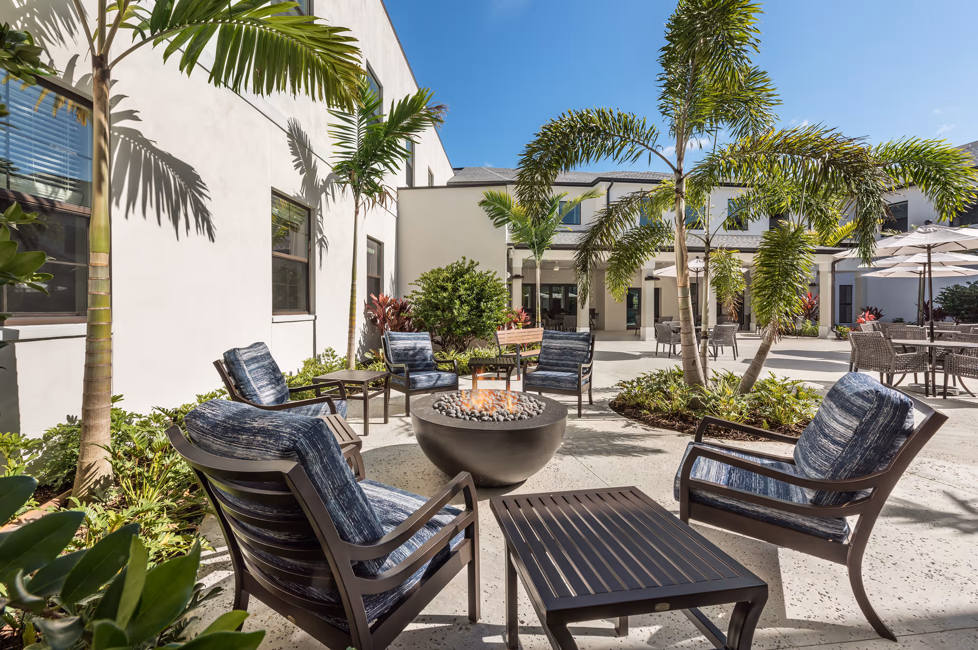 Outdoor patio area at HarborChase Of Stuart featuring cushioned chairs arranged around a modern fire pit, surrounded by palm trees and greenery under a clear blue sky.