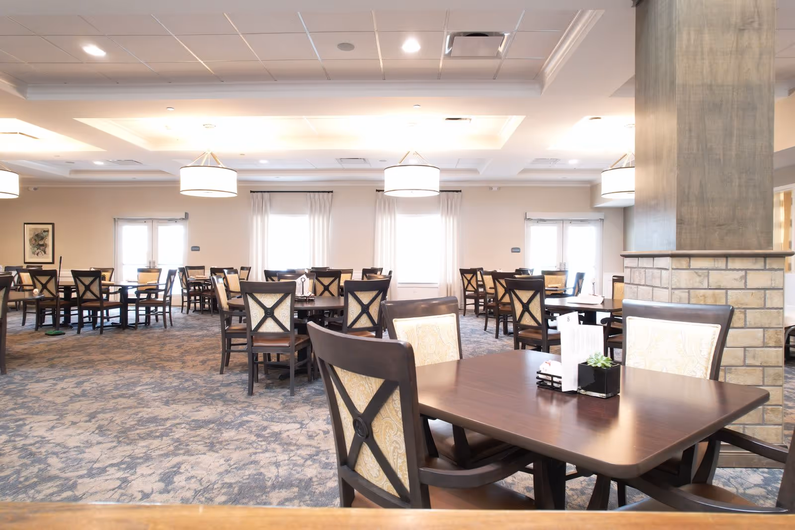 Bright, spacious dining room with multiple wooden tables and chairs, pendant lights, and a carpeted floor.