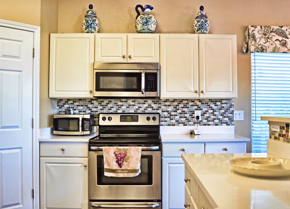 A modern kitchen with white cabinets, a stainless steel stove and microwave, a small black microwave on the counter, and a tiled backsplash in shades of gray and brown. Decorative ceramic jars are placed on top of the cabinets, and a window with blinds and a floral valance is visible on the right side.