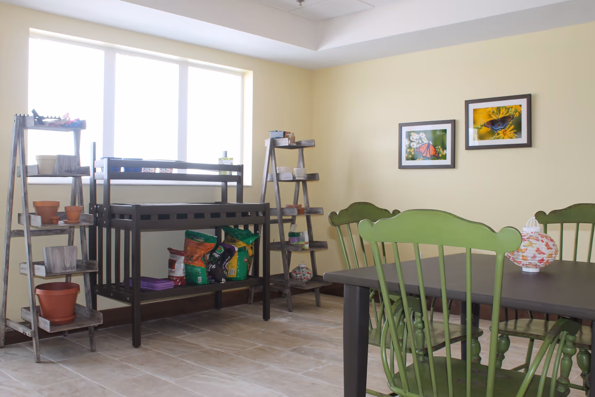 A bright room with a large window, featuring two wooden ladder-style shelves holding plant pots and gardening supplies. A dark wooden potting bench with bags of soil or fertilizer is positioned between the shelves. In the foreground, there is a dark table surrounded by green wooden chairs. Two framed pictures of butterflies on flowers hang on the light yellow wall.
