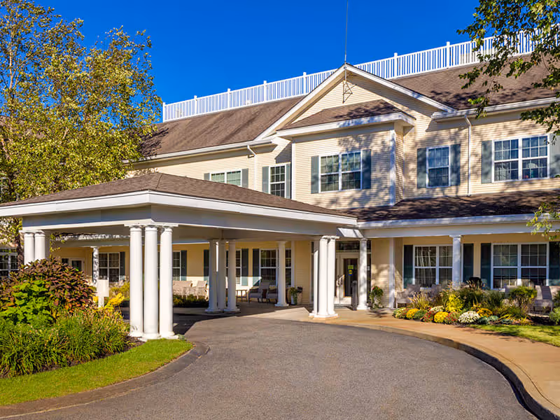 Exterior view of a senior living facility building with a covered entrance supported by white columns, surrounded by landscaped greenery and trees under a clear blue sky.