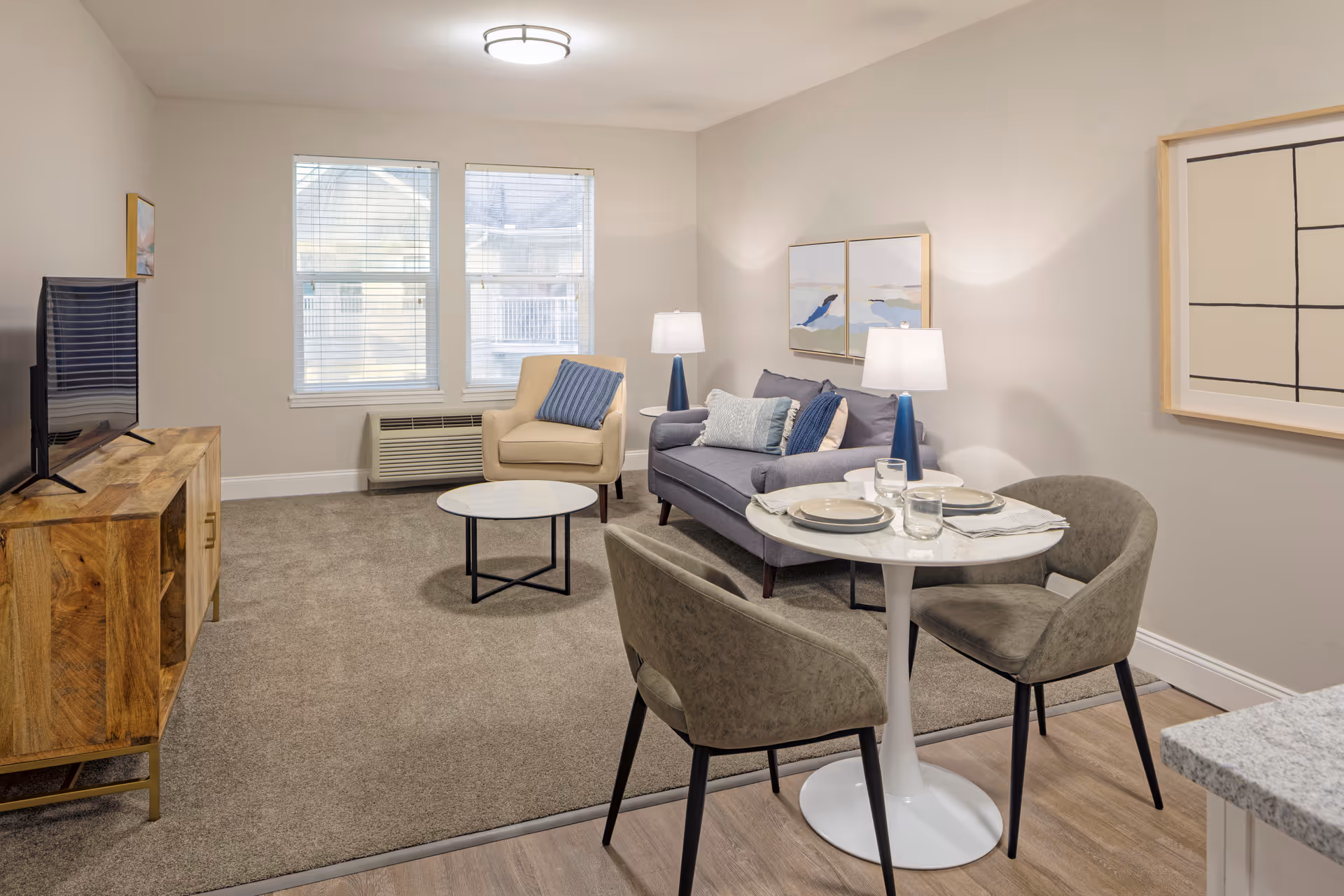 A cozy living room and dining area in a senior living facility. The living room features a gray sofa with blue and gray pillows, a beige armchair, two table lamps with blue bases, a round white coffee table, and a wooden TV stand with a flat-screen TV. The dining area has a small round white table set with plates and glasses, surrounded by two gray upholstered chairs. The room has beige walls, carpeted flooring in the living area, and wood flooring in the dining area. Two windows with blinds allow natural light to enter the space. Abstract artwork hangs on the walls.