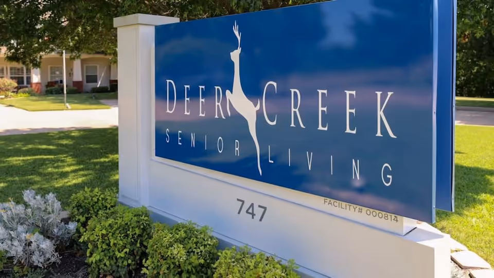 A large blue and white outdoor sign for Deer Creek Senior Living with a silhouette of a deer in the center, surrounded by green bushes and grass, with part of a building visible in the background.