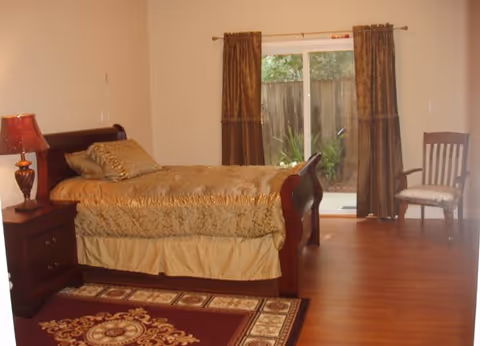 A bedroom with a wooden sleigh bed dressed in gold bedding, a nightstand with a lamp, a patterned area rug, sliding glass doors with curtains, and a chair.