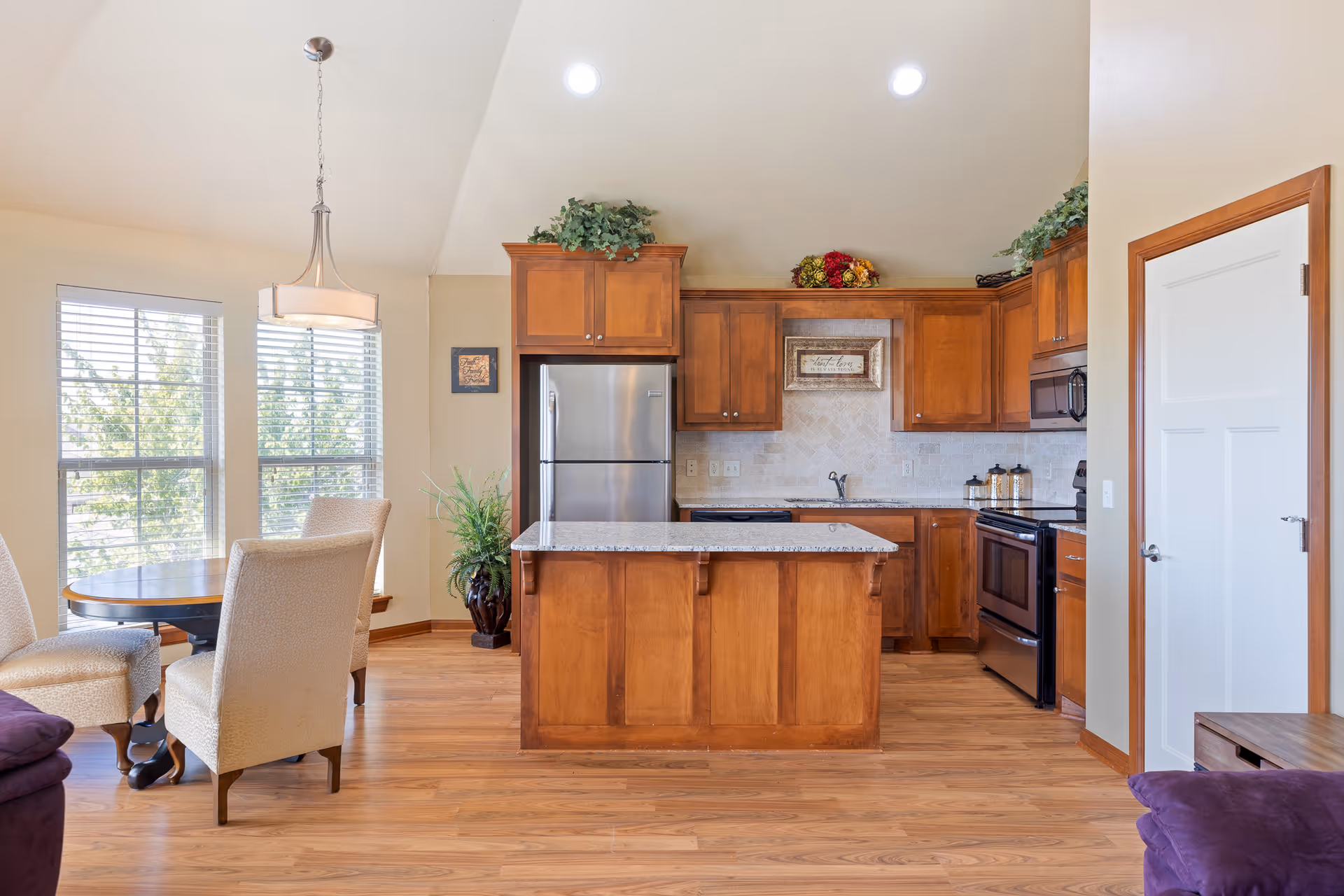 Bright open kitchen with a central island, wooden cabinets, stainless refrigerator, and an adjacent dining nook by large windows.