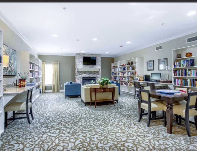 A spacious and well-lit common area with patterned carpet, bookshelves filled with books along the walls, a stone fireplace with a TV mounted above it, several chairs and tables including a desk with a computer, and a sofa set in the center with a small table holding a plant.