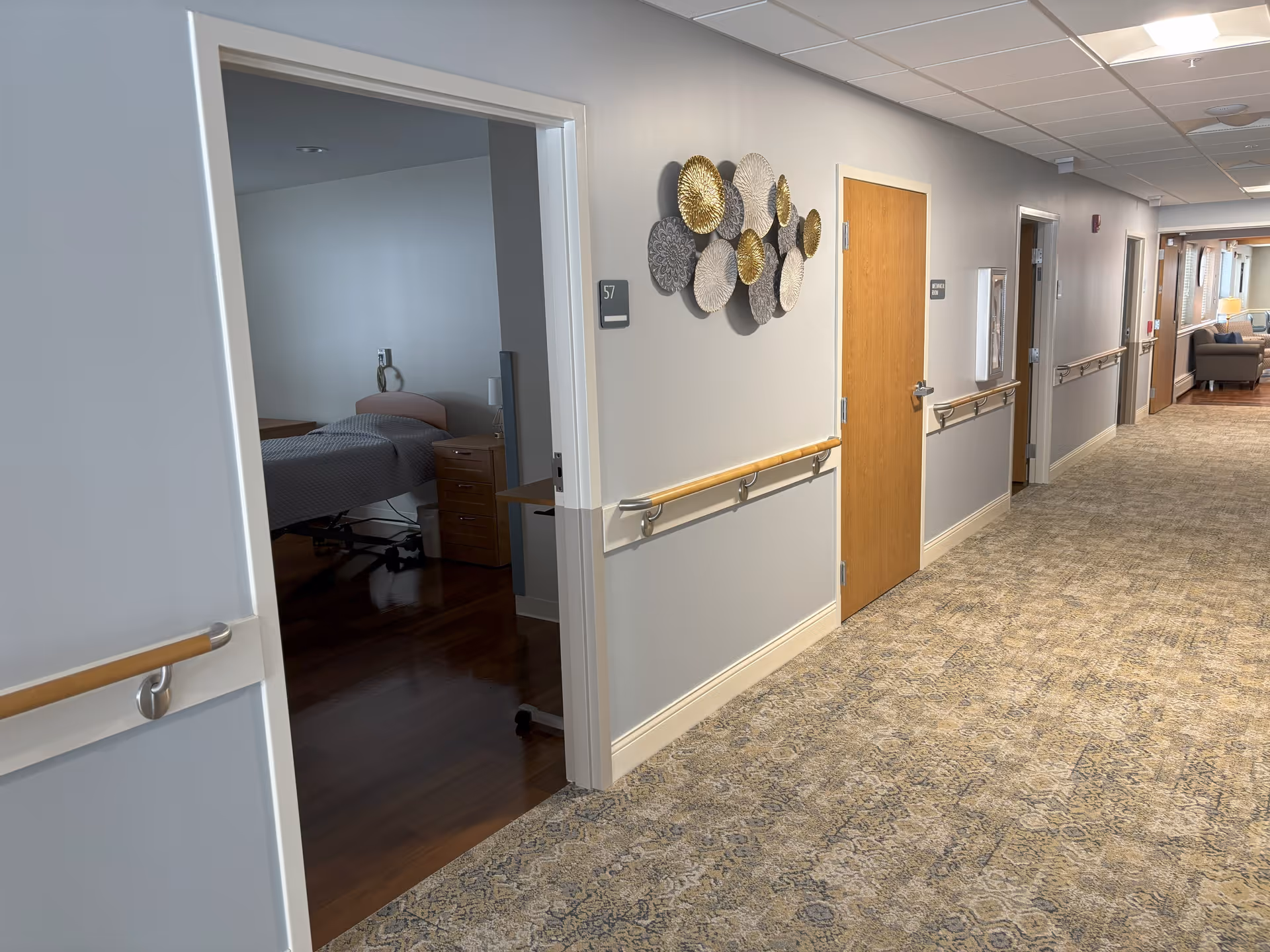 Carpeted hallway of a senior living facility with handrails, room doors, wall decor and an open bedroom visible to the left.