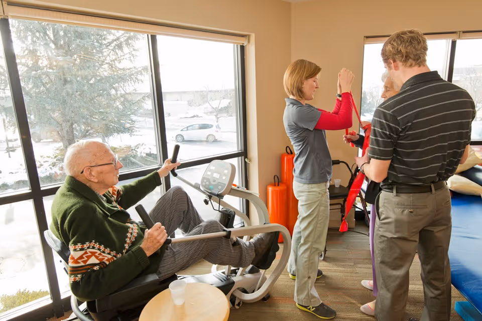 An elderly man exercises on a seated pedal machine near large windows with a snowy outdoor view. Two caregivers assist an elderly woman with a red resistance band in a bright room with exercise mats and equipment.