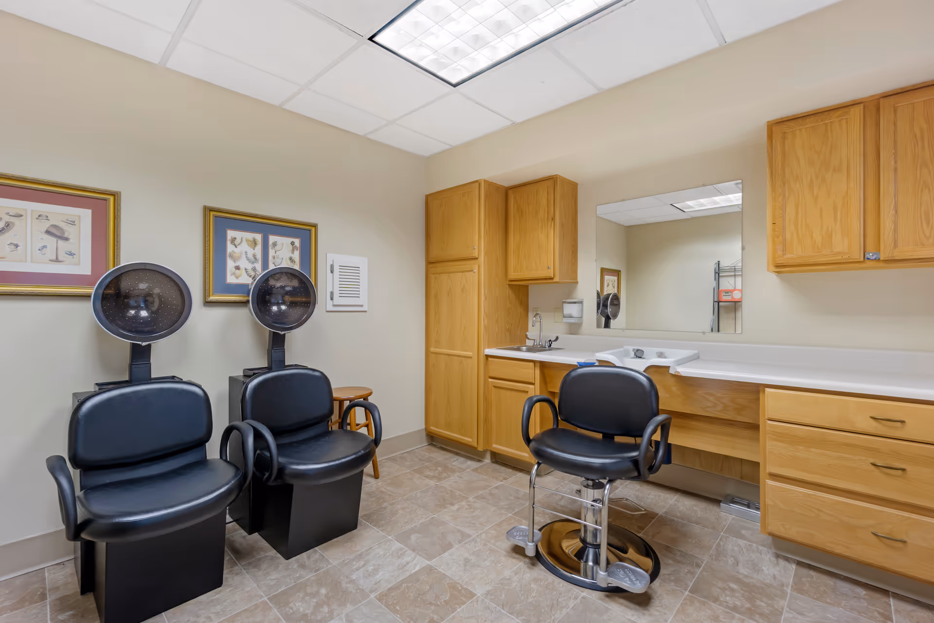 Interior of a salon room with two black hair drying chairs on the left and one black styling chair in front of a counter with a sink and large mirror. The room has beige walls, tiled floor, wooden cabinets, and framed pictures on the wall.