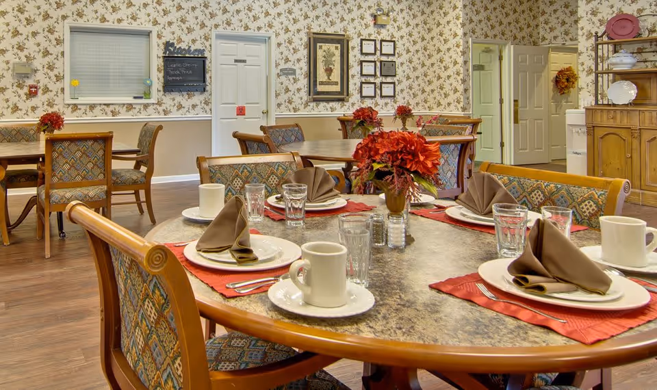 A dining room in an assisted living facility with several tables set for a meal. Each table has plates, cups, glasses, silverware, and brown folded napkins on red placemats. There are floral centerpieces on the tables. The room has patterned wallpaper, wooden chairs with patterned upholstery, and a wooden cabinet with dishes on the right side. Doors and framed pictures are visible on the walls.