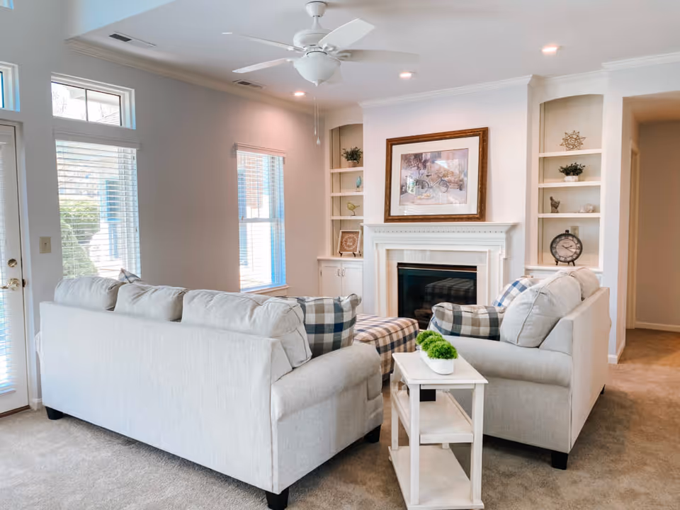 A bright living room with two light gray sofas facing each other, a plaid ottoman in the center, and a white side table with a small green plant. The room features a white fireplace with a framed picture above it, built-in shelves with decorative items, a ceiling fan, and large windows with blinds letting in natural light.