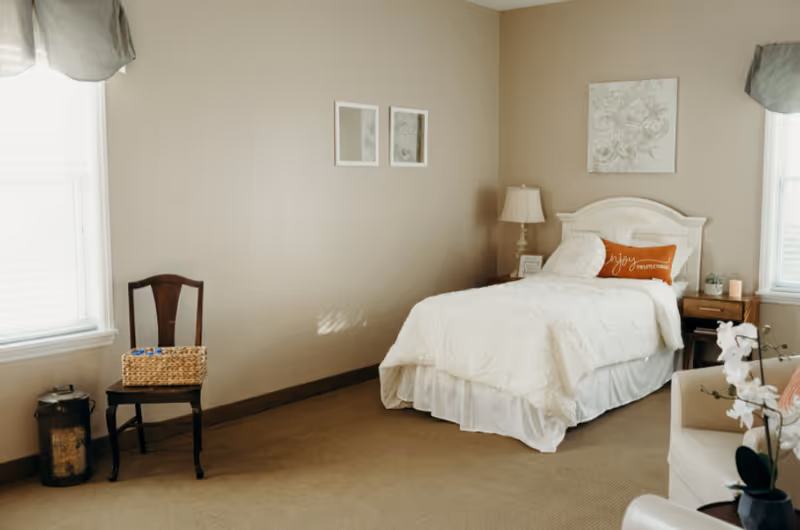 Sunny, neatly furnished bedroom with a single white bed, nightstand, chair, and windows.