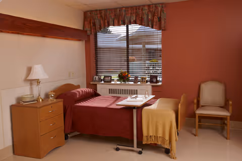 A small bedroom in a senior living facility with a single bed covered in a red bedspread and a yellow blanket at the foot. Next to the bed is a wooden nightstand with a white lamp and some items on it. A window with blinds and a floral valance is above a radiator, with several framed photos and a flower arrangement on the windowsill. A wooden chair with a cushioned seat and backrest is placed against the wall.