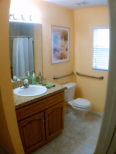 A small bathroom with yellow walls featuring a white sink with a granite countertop and wooden cabinet below. Above the sink is a large mirror with three light bulbs. To the right of the sink is a white toilet with metal grab bars on the walls behind and beside it. A framed picture hangs on the wall above the toilet, and a window with blinds is on the adjacent wall. The floor is tiled in a light color.