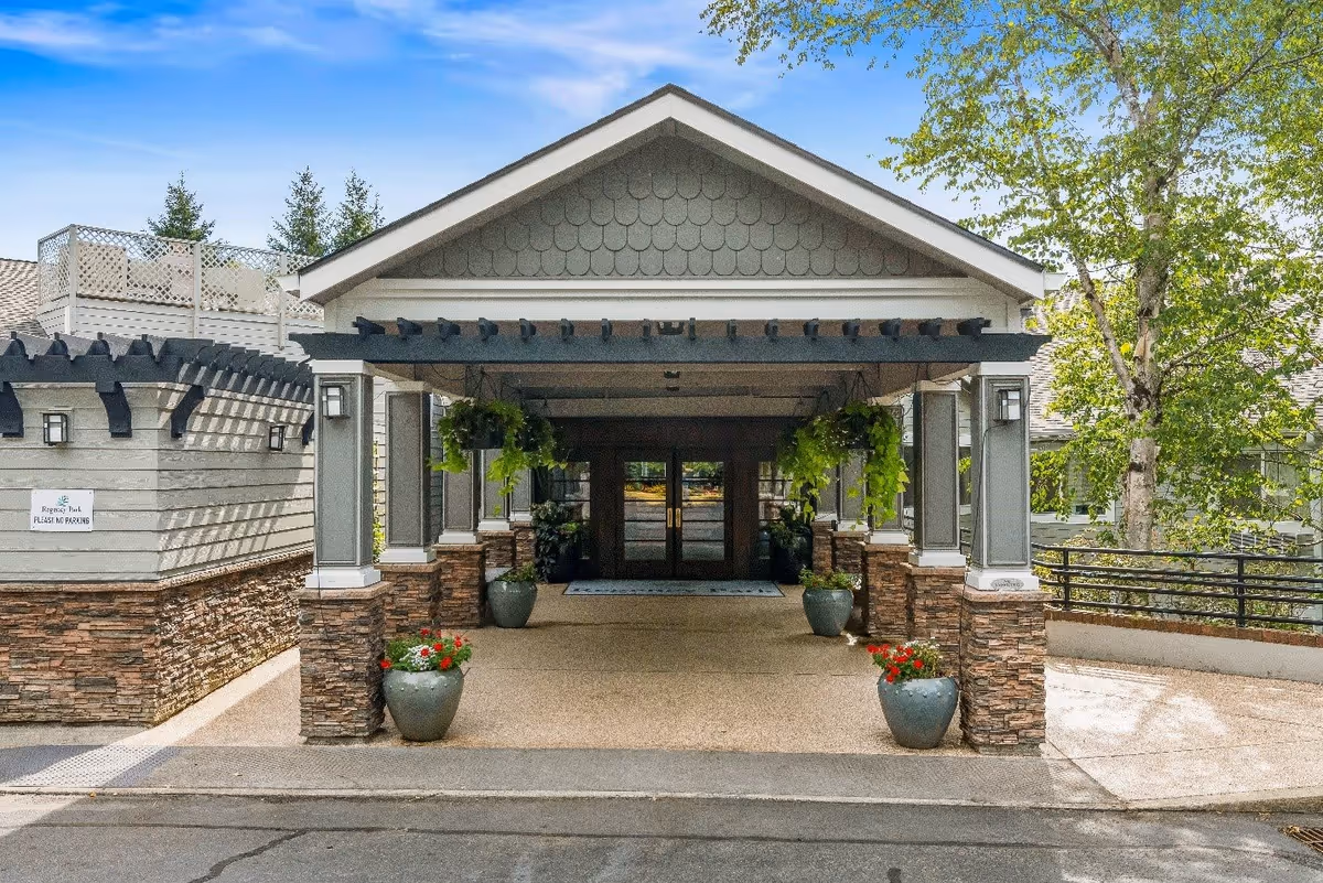 Entrance to a senior living facility with a covered driveway supported by stone and gray pillars, hanging plants, potted flowers, and double glass doors under a peaked roof. Trees and a clear blue sky are visible in the background.