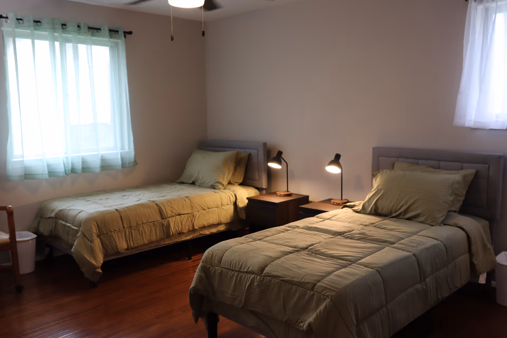 A simple bedroom with two single beds, each with green bedding and pillows. Between the beds are two dark wooden nightstands, each with a small lamp turned on. The room has wooden flooring and two windows with sheer curtains allowing natural light to enter.