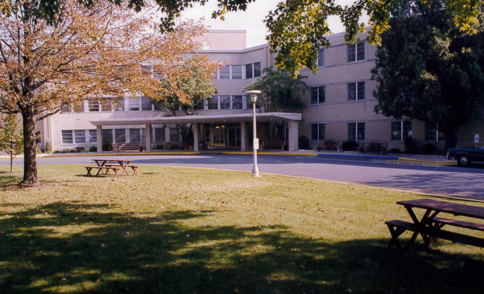 Front exterior of a three-story healthcare building with a covered entrance, driveway, lawn with picnic tables, and trees.
