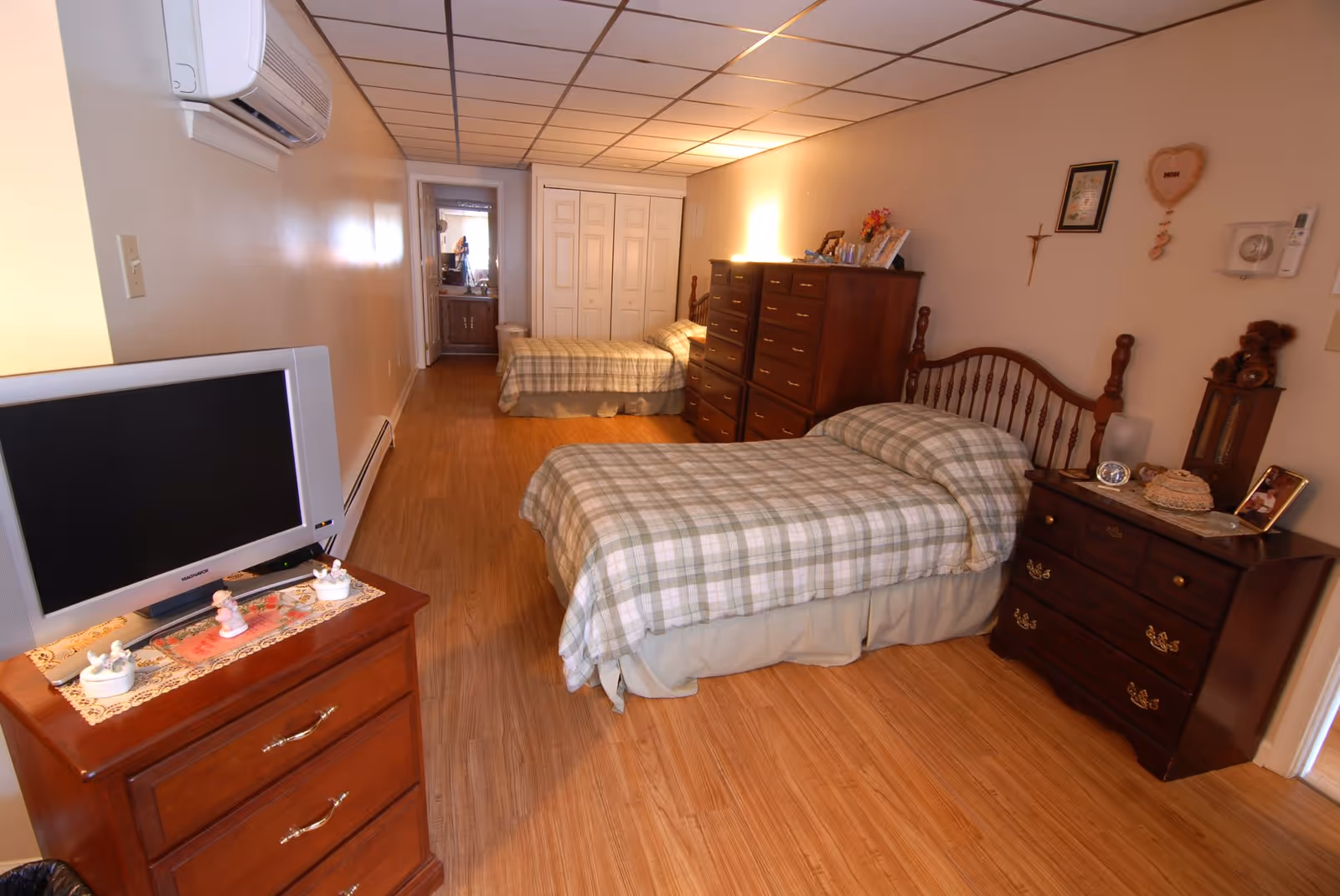 A senior living facility bedroom with two twin beds covered in matching plaid bedspreads. The room features wooden dressers, a TV on a small wooden cabinet, and various decorative items on the furniture. The floor is wooden, and there is an air conditioning unit mounted on the wall. A bathroom is visible through an open door at the far end of the room.