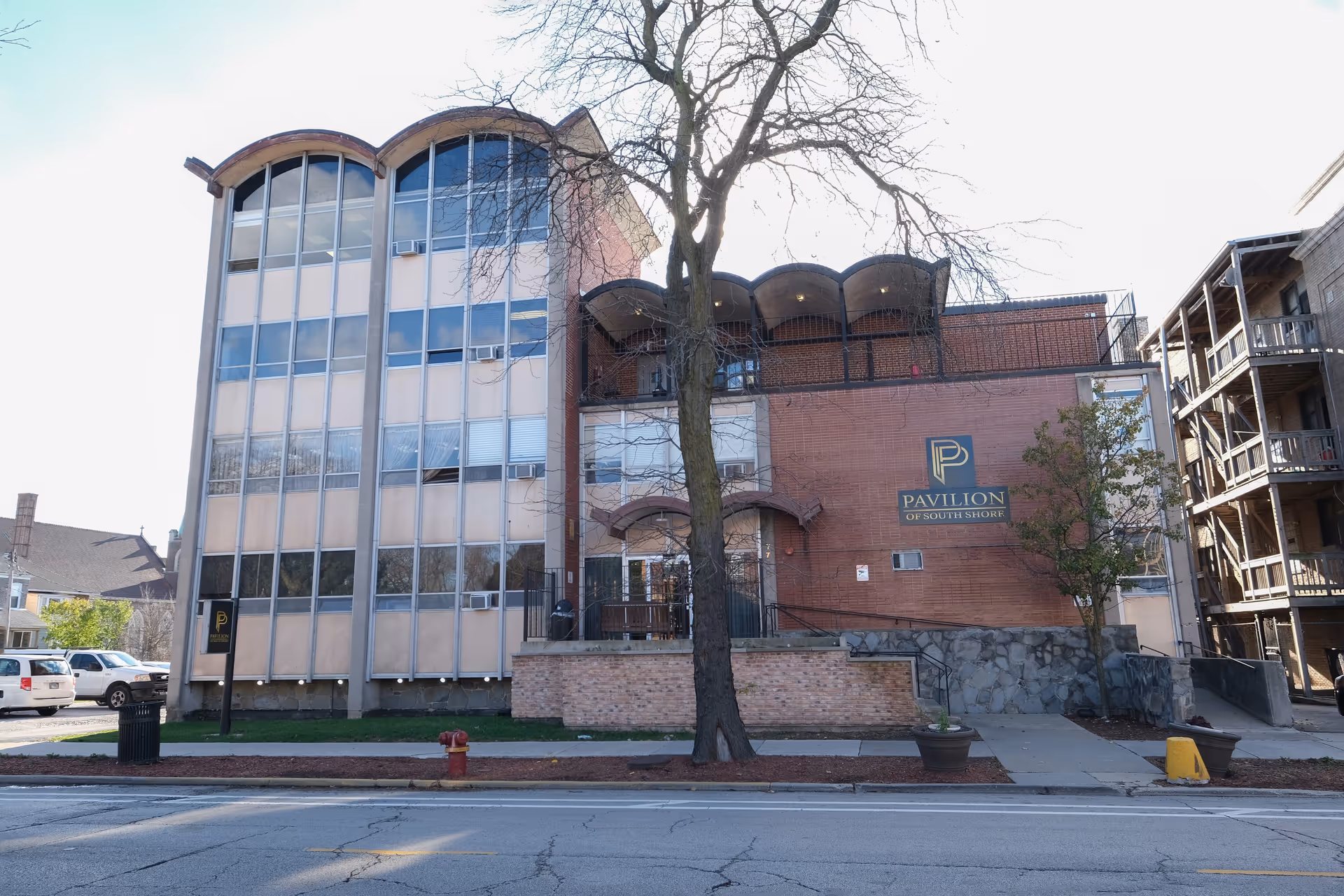 Exterior view of Pavilion of South Shore, a multi-story brick and glass building with a tree in front and a sign displaying the facility's name on the wall.