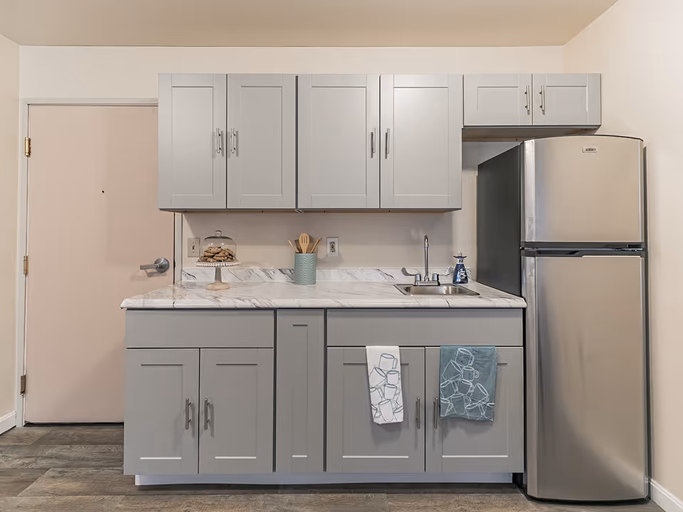 A small kitchen area with gray cabinets and drawers, a marble countertop, a stainless steel sink with a faucet, and a stainless steel refrigerator. There are two kitchen towels hanging on the cabinet doors, a container with wooden utensils, a soap dispenser, and a glass cake stand with cookies on the countertop. A closed door is visible to the left of the kitchen.