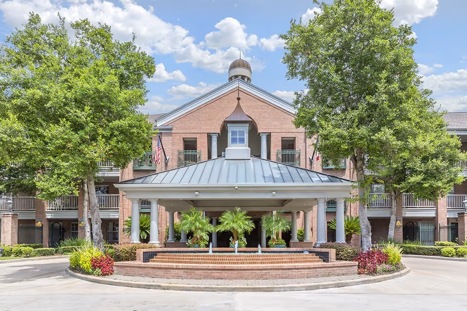 Front exterior view of a multi-story brick building with a covered entrance supported by columns, surrounded by trees and landscaping under a partly cloudy sky.