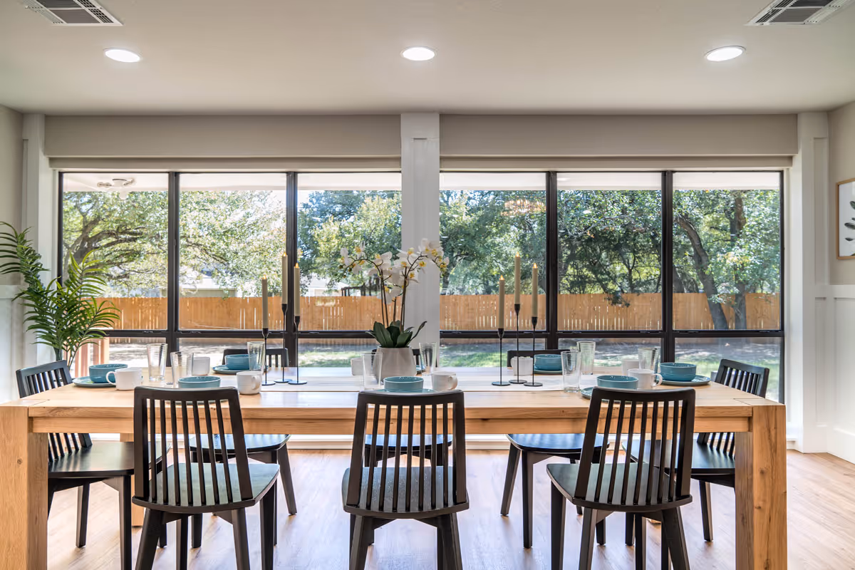 Bright dining room with a wooden table set for six and large windows overlooking a fenced backyard.