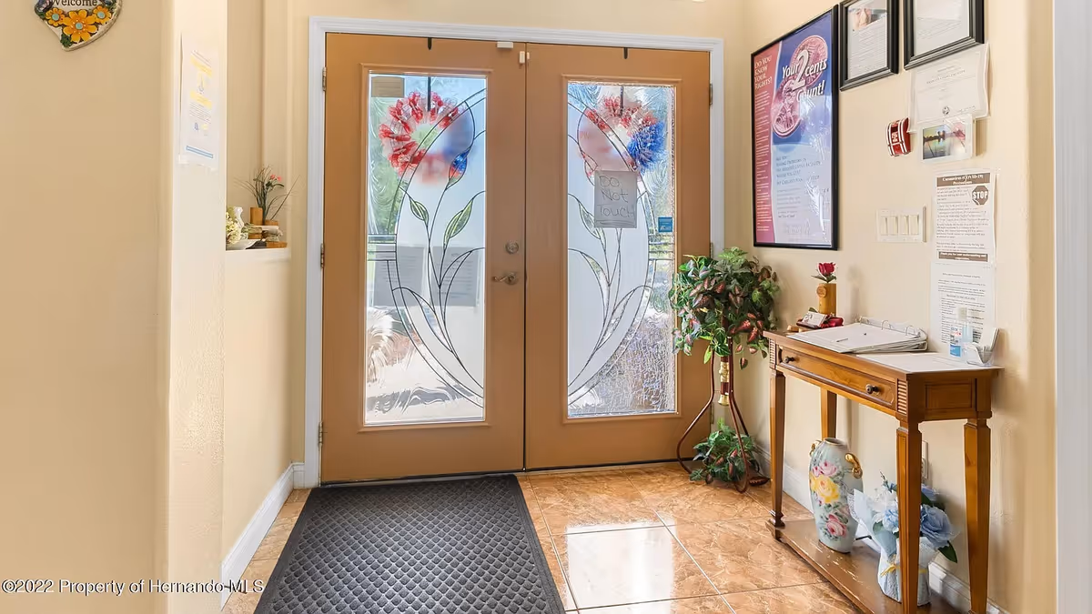 Entrance area of Aliviya Rose Manor Assisted Living Facility featuring double wooden doors with decorative frosted glass panels and floral designs. A small wooden table with a drawer is placed against the right wall, holding papers, a binder, a small vase with a red flower, and a hand sanitizer dispenser. Below the table are two decorative vases and a blue flower arrangement. A green potted plant stands next to the table. Various informational posters and notices are displayed on the wall above the table. The floor is tiled with a brownish pattern, and a black rubber mat is placed in front of the doors.