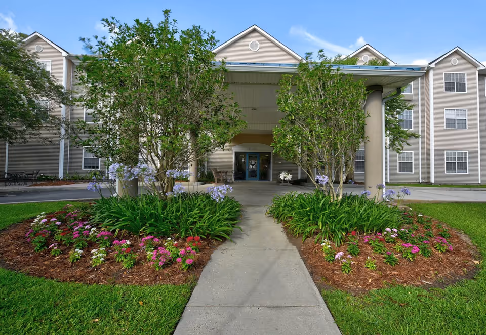Front entrance of a senior living facility with a covered walkway, flanked by two landscaped flower beds with green shrubs and colorful flowers. The building is beige with multiple windows and a blue sky with some clouds in the background.