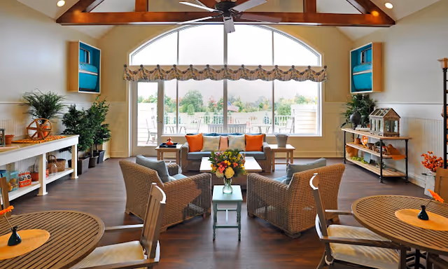 A bright and spacious living room area with high vaulted ceilings and exposed wooden beams. The room features a large arched window with a valance, allowing natural light to fill the space. There are two wicker armchairs and a sofa with orange and beige cushions arranged around a coffee table with a flower vase. Additional seating includes round wooden tables with chairs. The room is decorated with plants, shelves, and various decorative items, creating a welcoming and cozy atmosphere.