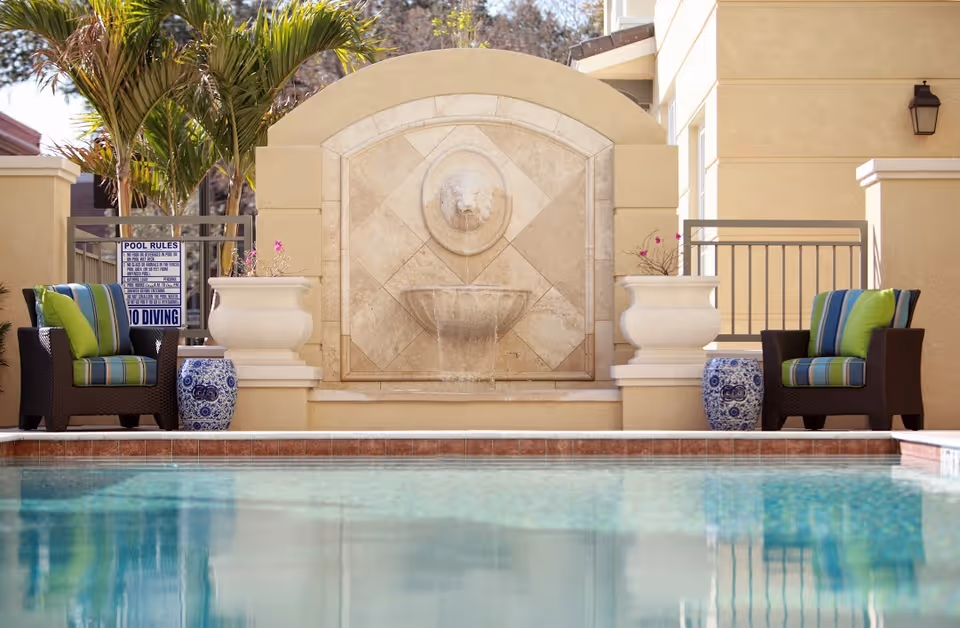Outdoor pool area featuring a decorative stone fountain, two cushioned lounge chairs with striped pillows, large planters, and the pool's reflection in the foreground.