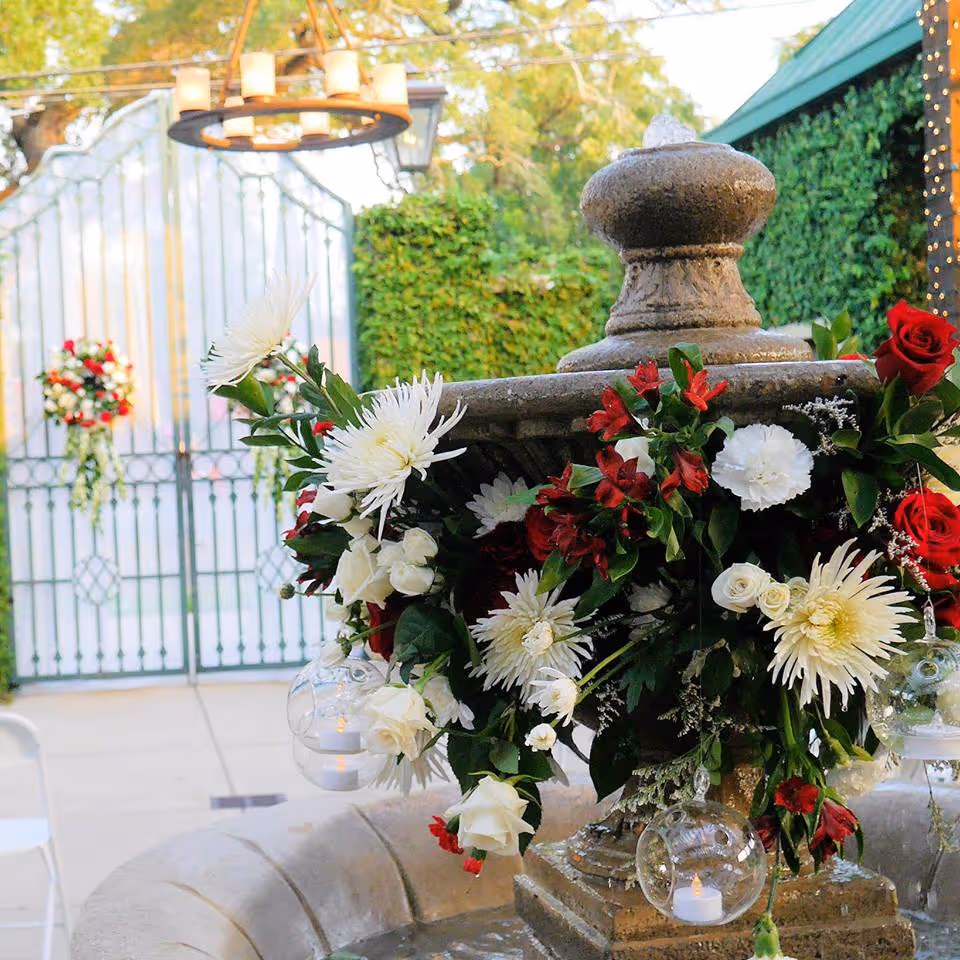 Stone fountain decorated with a floral arrangement of white and red flowers, including roses and chrysanthemums, with small glass candle holders hanging from the arrangement. In the background, there is a white metal gate adorned with a similar floral decoration and greenery-covered walls.